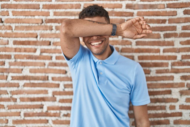 Brazilian young man standing over brick wall covering eyes with arm smiling cheerful and funny. blind concept.  - Photo, Image