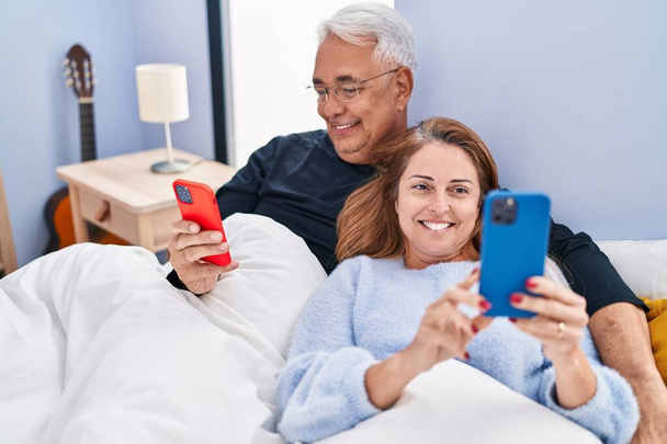 Middle age man and woman couple using smartphone lying on bed at bedroom - Foto, imagen