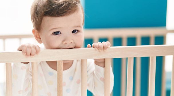 Adorable bebé caucásico de pie en la cuna mordiendo balaustrada de madera en el dormitorio - Foto, imagen