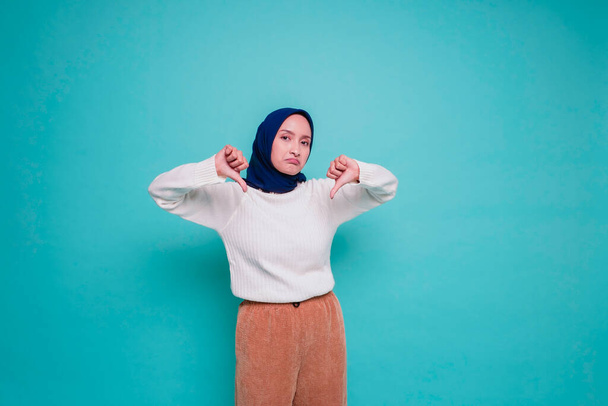 Disappointed Asian Muslim woman wearing white shirt and hijab gives thumbs down hand gesture of approval, isolated by blue background - Foto, Bild