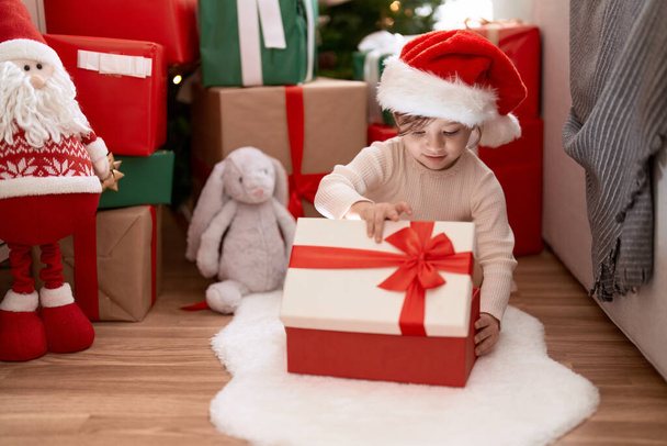 Очаровательная девушка распаковывает подарок сидя у рождественской елки дома - Фото, изображение