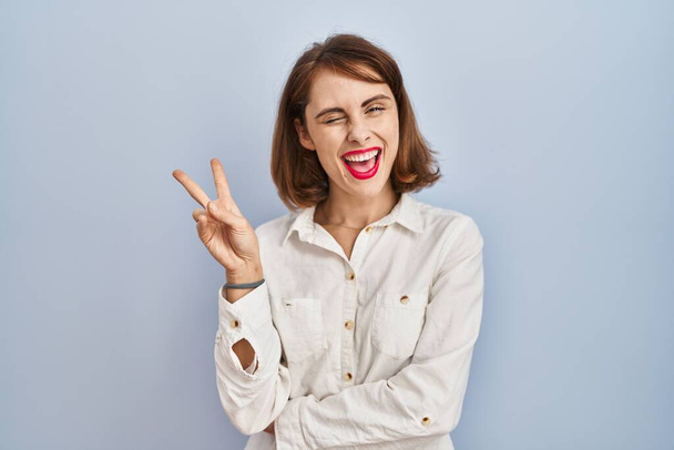Young beautiful woman standing casual over blue background smiling with happy face winking at the camera doing victory sign. number two.  - Photo, Image