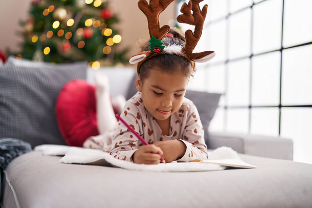 Adorable chica hispana dibujando en un cuaderno tumbada en un sofá junto al árbol de Navidad en casa - Foto, imagen