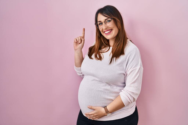 Mujer embarazada de pie sobre fondo rosa mostrando y apuntando hacia arriba con el dedo número uno mientras sonríe confiado y feliz.  - Foto, imagen