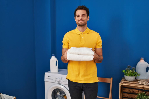 洗濯室できれいなタオルを持っている若いヒスパニック系男性が顔に深刻な表情でリラックスしました。カメラを見ているだけで.  - 写真・画像