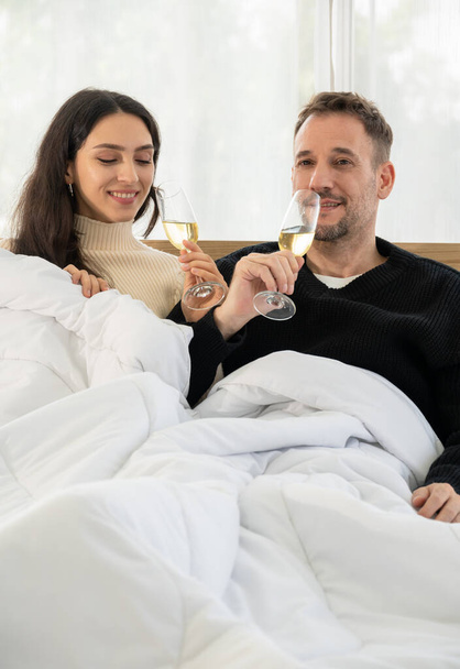 Romantic smiling couple lovers sitting on white bed and celebrating with glass of champagne in bedroom at home. Christmas, New Year, celebration  and good wishes concept - Φωτογραφία, εικόνα