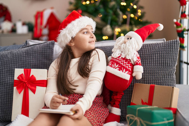 Adorabile ragazza ispanica che scrive sul taccuino seduto sul divano vicino all'albero di Natale a casa - Foto, immagini