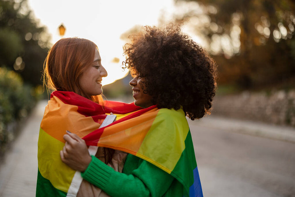 Multiracial pareja de lesbianas se miran con amor abrazándose con la bandera del arco iris lgbt en la calle al atardecer - Foto, Imagen