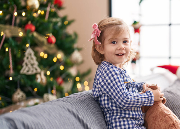 Adorable hispanic girl holding toy standing on sofa by christmas tree at home - Photo, image