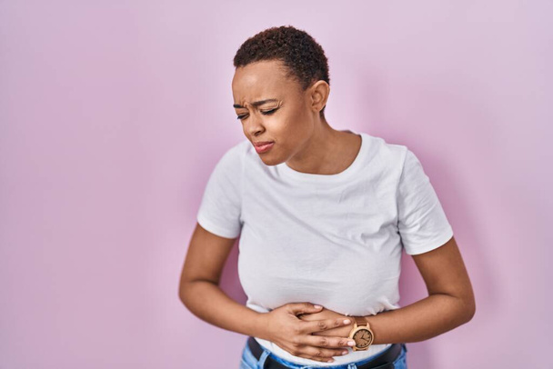 消化不良、痛みを伴う病気が体調不良のため、美しいアフリカ系アメリカ人女性が胃の上に手でピンクの背景に立っている。キャッシュ・コンセプト.  - 写真・画像