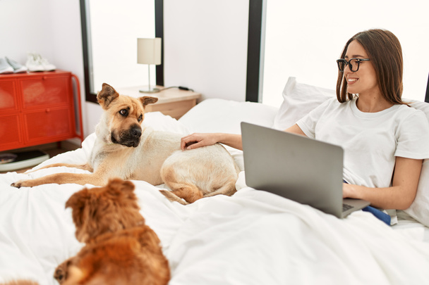 Junge hispanische Frau sitzt mit Laptop im Bett mit Hunden im Schlafzimmer - Foto, Bild