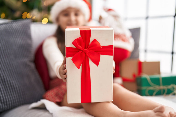 Adorabile ragazza ispanica che tiene il regalo seduto sul divano vicino all'albero di Natale a casa - Foto, immagini