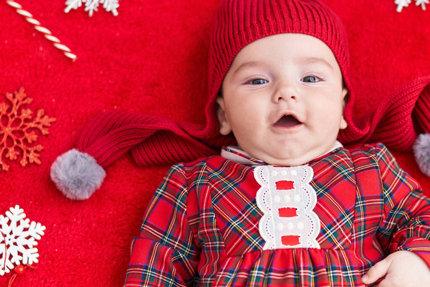 Liebenswertes kaukasisches Baby auf dem Boden liegend mit Weihnachtsdekor über isoliertem rotem Hintergrund - Foto, Bild