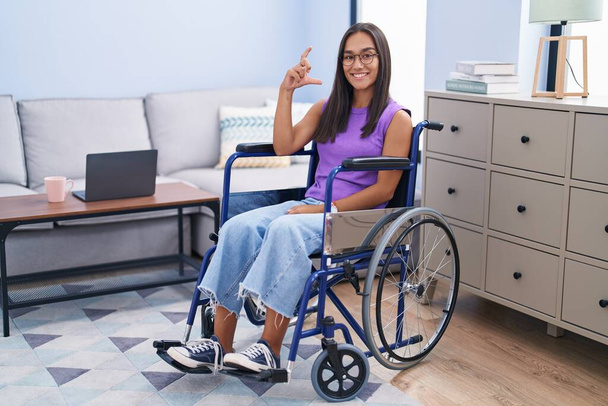 Młoda Latynoska kobieta siedząca na wózku inwalidzkim w domu uśmiechnięta i pewna siebie gestykulująca ręką robiąc mały znak z palcami patrzącymi i kamerą. Koncepcja środka.  - Zdjęcie, obraz
