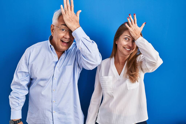 Couple hispanique moyen âge debout sur fond bleu surpris avec la main sur la tête pour erreur, rappelez-vous erreur. oublié, mauvais concept de mémoire.  - Photo, image