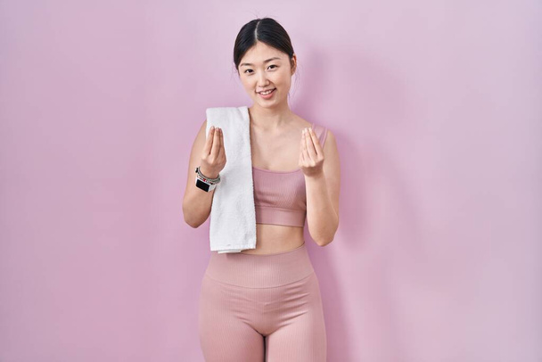 Mujer joven china con ropa deportiva y toalla haciendo gesto de dinero con las manos, pidiendo pago de sueldo, negocio millonario  - Foto, imagen