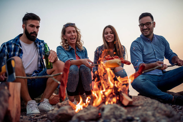 日没時に火の周りに座っている友人のグループ,ソーセージを焼くとビーチで素晴らしい時間を過ごして..  - 写真・画像