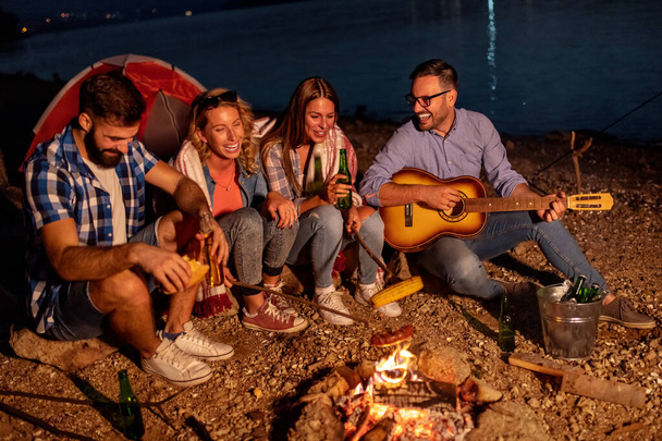 Eine Gruppe von Freunden sitzt nachts am Feuer und hört dem Gitarristen zu, grillt Würstchen und amüsiert sich am Strand.  - Foto, Bild
