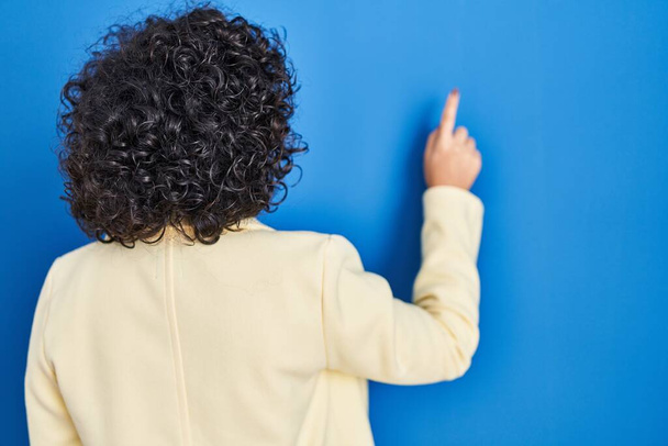 Young brunette woman with curly hair standing over blue background posing backwards pointing ahead with finger hand  - Photo, image