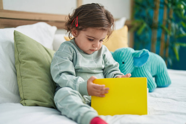 Adorable hispanic girl reading book sitting on bed at bedroom - Photo, Image
