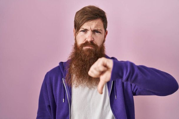 Кавказький чоловік з довгою бородою, що стоїть над рожевим фоном, виглядає нещасним і сердитим, виявляючи відмову і негативний з пальцями вниз. поганий вираз.  - Фото, зображення