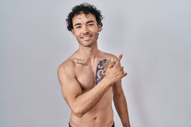 Hombre hispano de pie sin camisa alegre con una sonrisa de la cara señalando con la mano y el dedo hacia el lado con expresión feliz y natural en la cara  - Foto, imagen