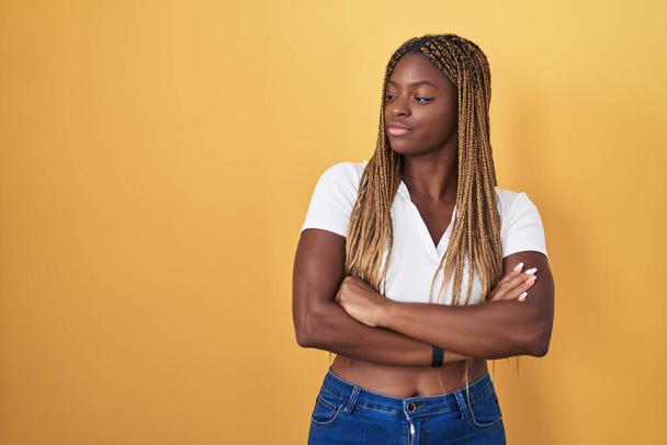 Afroamerykanka z plecionymi włosami stojąca nad żółtym tłem patrząca z boku z ramionami skrzyżowanymi przekonana i pewna siebie  - Zdjęcie, obraz