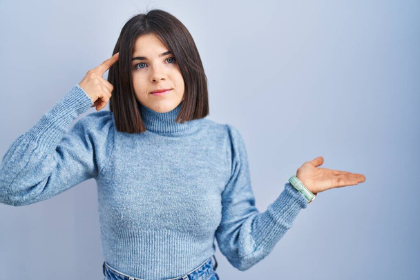 Jonge Spaanse vrouw die over een blauwe achtergrond staat verward en geïrriteerd met een open handpalm die kopieerruimte toont en met de vinger naar het voorhoofd wijst. Denk er eens over na..  - Foto, afbeelding