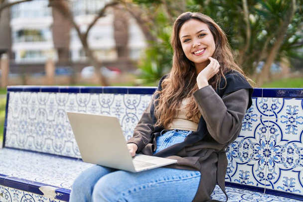 Young beautiful hispanic woman using laptop sitting on bench at park - Foto, afbeelding
