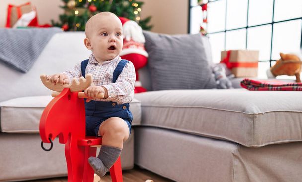 Adorable caucasian baby playing with reindeer rocking by christmas tree at home - Photo, Image