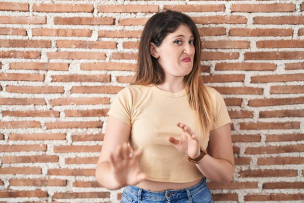 Young brunette woman standing over bricks wall disgusted expression, displeased and fearful doing disgust face because aversion reaction.  - Photo, Image