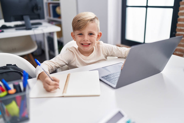 Αξιολάτρευτος μαθητής μικρών παιδιών που χρησιμοποιεί το laptop γράφοντας στο σημειωματάριο στην τάξη - Φωτογραφία, εικόνα