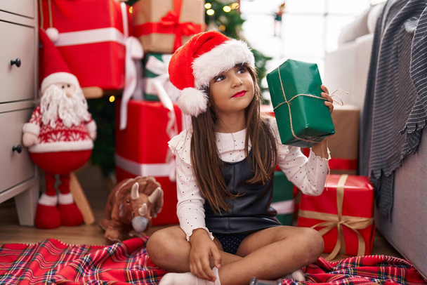 Чарівна іспаномовна дівчина тримає подарунок, сидячи на підлозі на ялинці вдома
 - Фото, зображення