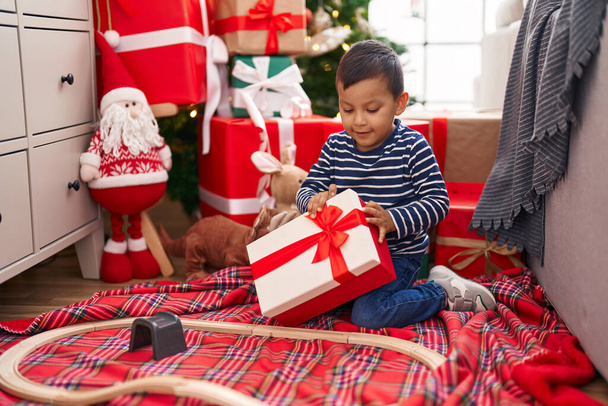 Adorable hispanic toddler holding gift sitting on floor by christmas tree at home - Photo, Image