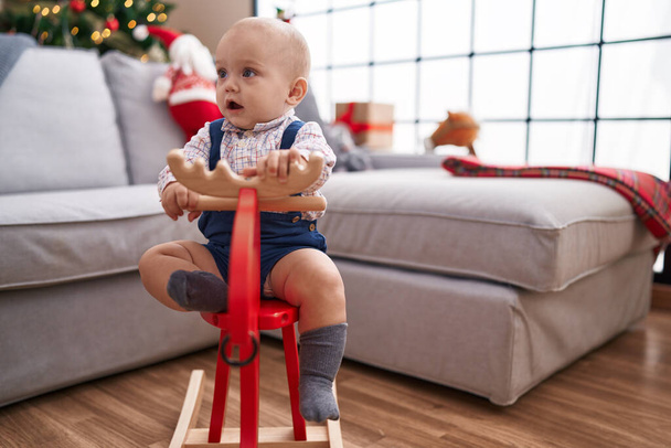 Adorable caucasian baby playing with reindeer rocking by christmas tree at home - Fotografie, Obrázek