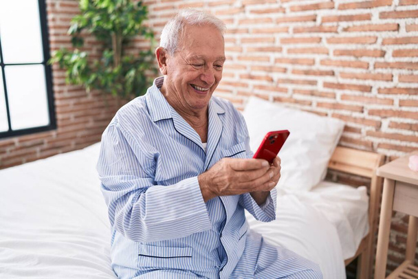 Middle age grey-haired man using smartphone sitting on bed at bedroom - Photo, Image