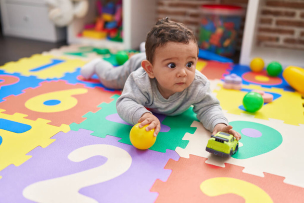 Αξιολάτρευτο ισπανόφωνο μωρό παίζει με μπάλα και αυτοκίνητο που βρίσκεται στο πάτωμα στο νηπιαγωγείο - Φωτογραφία, εικόνα