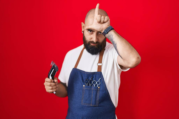 Молодий іспаномовний чоловік з бородою та татуюваннями носить бар'єрний фартух, який тримає бритву, насміхаючись над людьми з пальцями на лобі, роблячи жест невдахою, глузуючи та ображаючи
.  - Фото, зображення