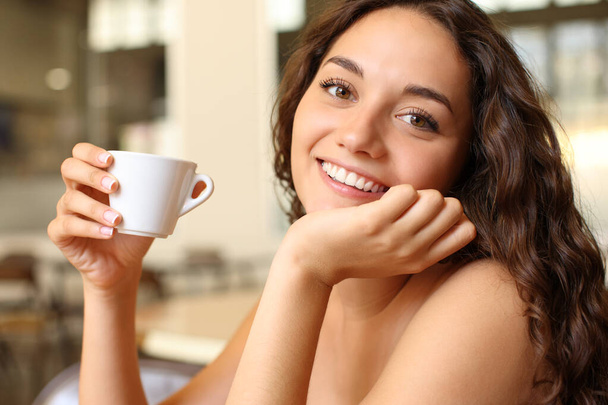 Happy woman holding coffee cup looks at you sitting in a bar - Фото, изображение