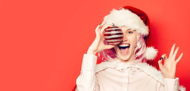 Funny winking woman with christmas ball. Pretty girl celebrate winter holidays on red isolated background Young beautiful girl with christmas ball - 写真・画像