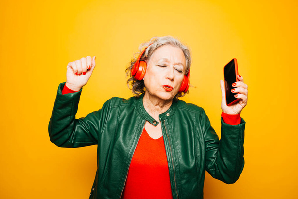Mujer mayor con auriculares rojos y smartphone rojo, bailando sobre un fondo amarillo - Foto, imagen