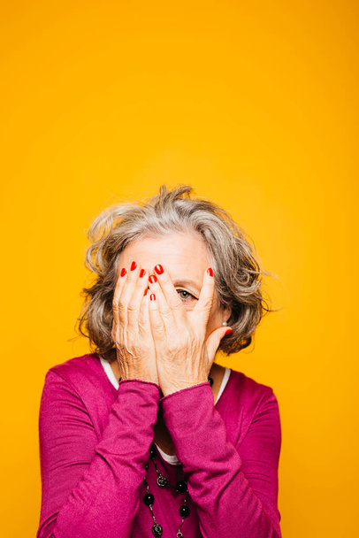 Porträt einer Seniorin, die ihr Gesicht mit den Händen vor gelbem Hintergrund bedeckt - Foto, Bild