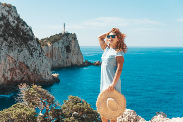 Девушка в синем платье стоит на холме рядом с белым маяком, наблюдая за морем внизу на берегу Лефкады. Греция - Фото, изображение