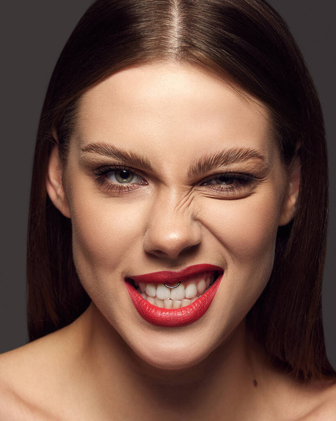 Close-up portrait of emotive beautiful young woman with bright makeup, grimacing over dark grey background. Mouth septum, piercing. Concept of beauty, fashion, makeup, magazine, emotions and ad. - Фото, зображення