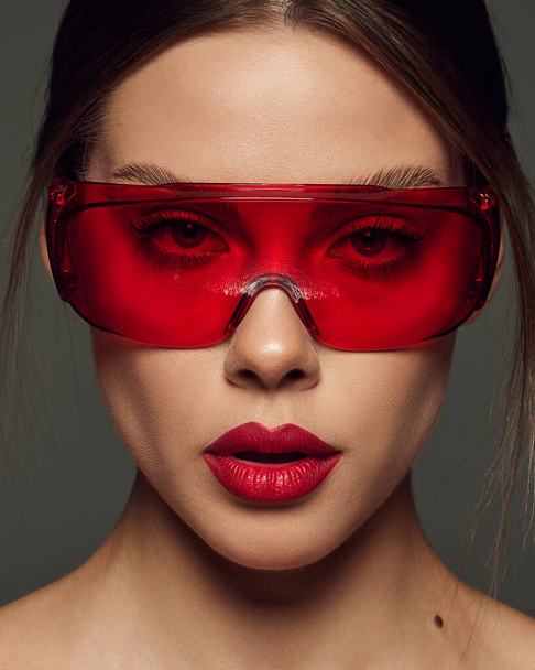 Close-up portrait of beautiful young woman with red lipstick posing in red plastic glasses on dark grey background. Cosmetology treatment. Concept of beauty, fashion, makeup, magazine, emotions and ad - Fotó, kép