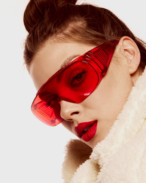 Close-up portrait of young with well-kept skin, trendy makeup and red glasses posing in furry coat isolated over white background. Concept of hight fashion, style, make-up, beauty, magazine style, ad - Фото, изображение