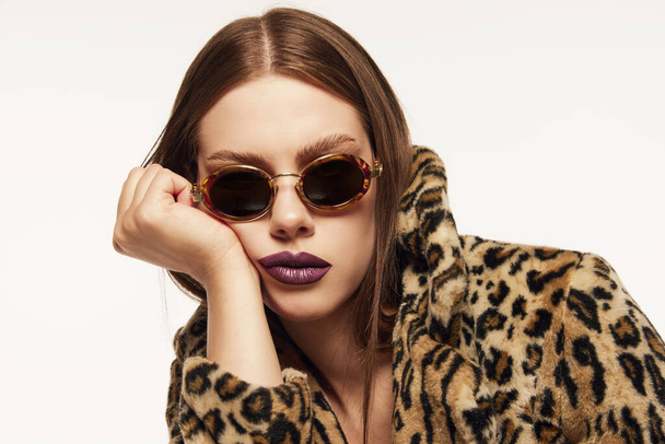Portrait of beautiful woman posing in trendy sunglasses and animal print coat over white background. Plum colored lipstick. Bored look. Concept of style, beauty, high fashion, magazine style and ad - Фото, изображение
