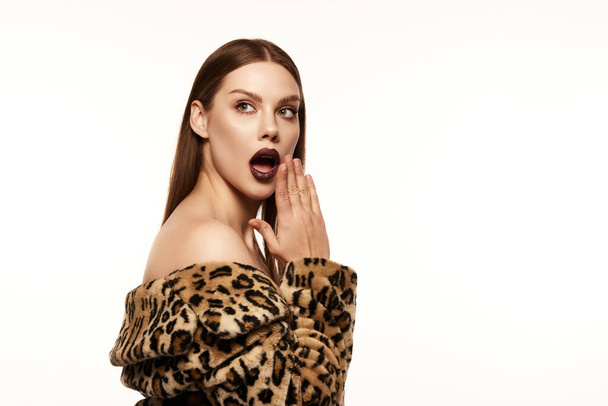 Portrait of beautiful woman posing in animal print coat with dark lips makeup look over white background. Shocked expression. Concept of style, emotions, beauty, high fashion, magazine style and ad - Фото, зображення