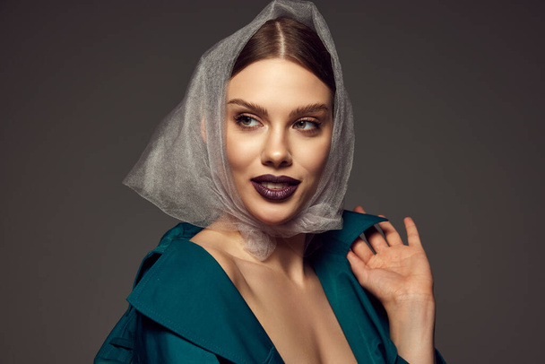 Portrait of young stylish woman with trendy dark lips make-up posing in fashionable coat and transparent headscarf over dark grey background. Stylish season. Concept of beauty, high fashion, magazine - Fotografie, Obrázek