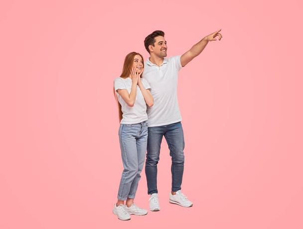 Full body happy young man in casual clothes smiling and pointing away for amazed girlfriend while standing against pink background together - Foto, immagini